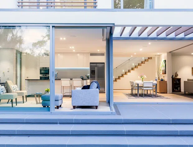 A bright contemporary two-storey home viewed from the exterior, with broad blue-framed sliding glass doors opening onto a stepped terrace. The open-plan interior visibly includes a modern kitchen, living area, dining area, and a timber stairway. White walls, pale blue upholstery, light timber stair treads, and exposed pergola beams create a clean, airy composition.