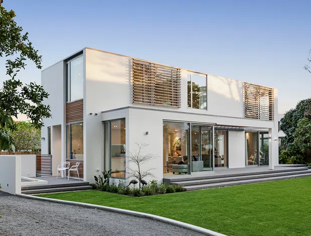 A contemporary two-storey white-rendered residence with a clean rectilinear form, extensive floor-to-ceiling glazing, timber louvres, and broad outdoor steps opening onto a manicured lawn. The suburban garden setting, flat roofline, and restrained landscaping reinforce the modern architectural character.