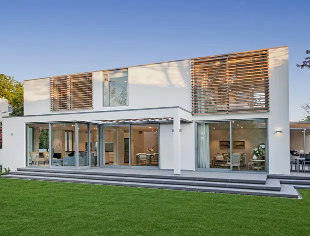 Two-storey contemporary white-rendered home photographed from the lawn, with extensive floor-to-ceiling glazing, deep upper-level timber louvres, a flat roofline, covered terrace, and broad stepped outdoor platform. Interior living and dining areas are visible through the glazing, with an outdoor dining area at the right.