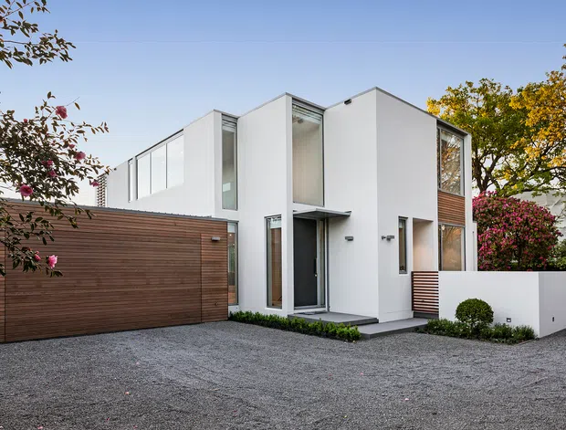 Photograph of a contemporary two-storey white rendered house with a strong geometric composition, tall glazing, warm horizontal timber cladding, and an attached timber-clad garage. The property is approached across a broad gravel driveway and softened by manicured shrubs and mature planting.