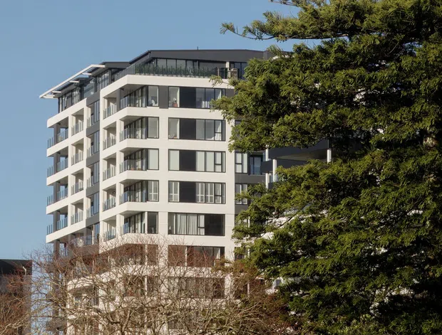 Exterior view of a contemporary multi-storey apartment building, partially screened by mature trees. The building has a light-coloured facade, extensive dark-framed glazing, projecting balconies, and a dark flat roof against a clear blue sky.