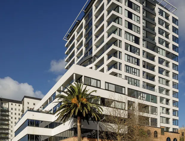 A tall contemporary multi-storey apartment building viewed from a street corner, with white geometric balconies, extensive dark-framed glazing, and a lower podium clad in warm brick with arched windows. The building rises above an urban streetscape under a clear blue sky, with a prominent palm tree in front.