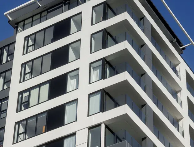 Upward-looking exterior photograph of a contemporary multi-storey apartment building. The facade is predominantly white with dark-framed floor-to-ceiling windows, projecting balconies, glass balustrades, and a distinctive flat roof canopy with projecting metal elements against a clear blue sky.