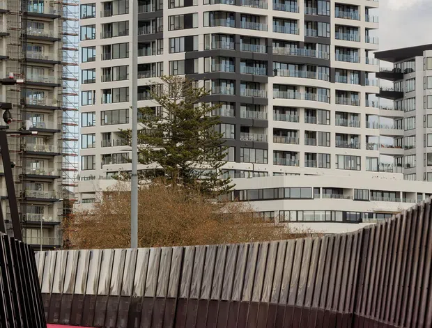An urban architectural exterior view showing a contemporary multi-storey apartment building with white and dark grey facade panels, extensive balconies, and large glazed windows. The foreground features a vivid pink pedestrian bridge or elevated walkway with dark vertical metal balustrades. Additional apartment construction and buildings are visible around it.