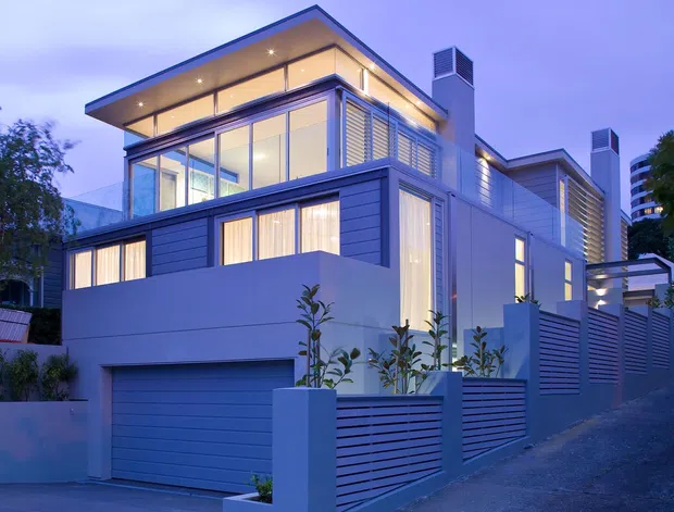 Twilight exterior view of a contemporary multi-level urban residence. The home features extensive floor-to-ceiling glazing, projecting flat roof planes, horizontal cladding, rendered walls, rooftop-style screening and integrated warm lighting. A garage, driveway, front fence and landscaped planters are visible in the foreground.
