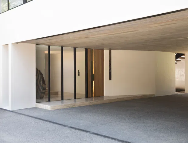 Minimalist modern covered entry or carport area with a long white rendered facade, timber-lined soffit, full-height black-framed glazing, and a timber pivot-style entry door. Pale stone paving defines the threshold, while the broad dark paved foreground suggests a driveway or sheltered vehicle area. The composition is clean and architectural, with strong horizontal lines and restrained natural materials.