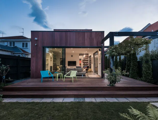 A compact contemporary backyard extension or pavilion with a dark timber-clad upper facade, large glazed opening, and a broad timber deck. The open interior reveals a living area with shelving and seating, while the landscaped garden, privacy fencing, neighbouring homes, and pitched rooflines frame the structure. The image is photographed from the exterior at dusk.