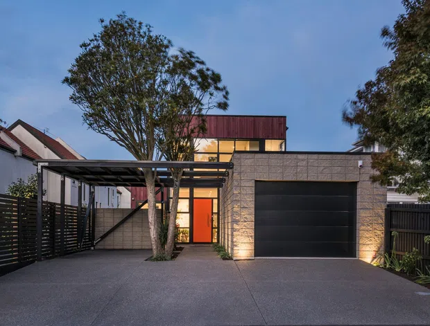 Street-facing contemporary residential facade at dusk, featuring a broad dark paved driveway, a black horizontal-slat carport or entry canopy, a large black sectional garage door, pale masonry walling, warm glazed entry lighting, and a raised upper volume with dark reddish cladding. Established trees and neighbouring pitched-roof homes frame the property.