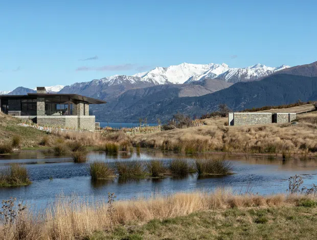 A low, contemporary single-storey house sits beside a natural pond in a dry tussock landscape. The building has dark horizontal cladding, extensive glazing, a broad low-pitched roof with deep eaves, and stone masonry walls. Snow-capped mountain ranges and a lake or broad water body are visible beyond the property.