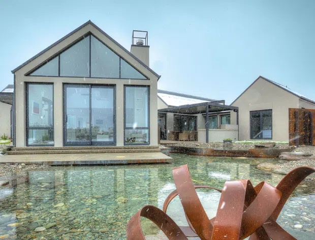 Exterior view of a contemporary gabled residence beside a shallow reflective pond or water feature. The main pavilion has a glazed gable façade, dark metal-framed windows, light rendered walls, and a projecting low roof edge. Additional connected gabled volumes and a covered outdoor area extend behind it, with timber screening on the right. Snow is visible around the landscaped site, and a sculptural rust-coloured metal feature occupies the foreground.