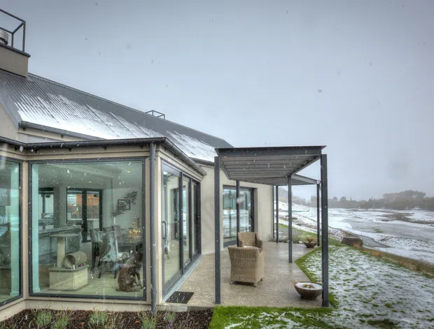 Exterior view of a contemporary glass-walled home beside a snowy waterfront or tidal landscape. The building features extensive floor-to-ceiling glazing, a corrugated metal pitched roof, a tall chimney, and a covered outdoor terrace with slim metal supports and wicker seating.
