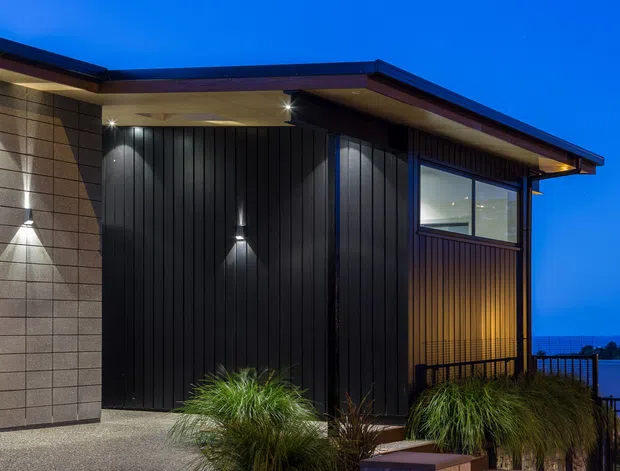 Twilight exterior view of a contemporary single-storey building with a low-pitched projecting roof, dark vertical cladding, textured light masonry wall panels, recessed exterior lighting, large corner glazing, and landscaped ornamental grasses. A balcony or deck railing and distant water or landscape are visible beyond the building.