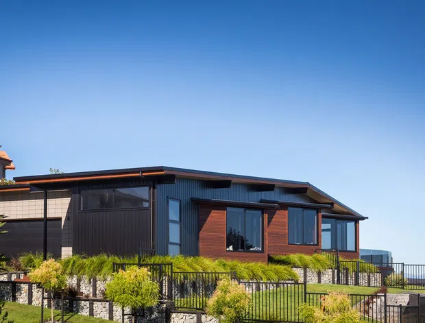 Single-storey contemporary home viewed from the exterior, with a low-pitched roof, dark vertical cladding, warm timber feature walls around the windows, extensive glazing, and a terraced landscaped site enclosed by black metal fencing. The house has a strong horizontal composition and modern residential detailing.