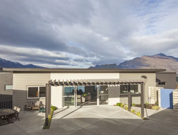 Exterior view of a contemporary single-storey home with a low-pitched roof, light horizontal cladding, dark grey rendered sections, extensive glazing, and a concrete courtyard or driveway. A timber pergola shades the front glazing, with mountain terrain visible in the background and a small outdoor seating area in the foreground.