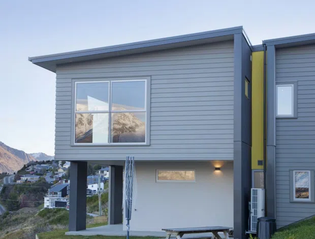 Exterior view of a contemporary multi-level home on a steep hillside, featuring grey horizontal cladding, simple projecting flat roof forms, large rectangular windows, and a bold lime-green vertical accent panel. The building is elevated above the sloping site, with mountain and suburban hillside views in the background.