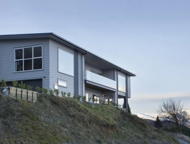 A contemporary two-storey residence perched on a sloped site, viewed from below. The building has horizontal grey weatherboard cladding, dark window and door frames, broad glazed openings, a projecting upper-level balcony or terrace, and a low-pitched roof. Mountain peaks are visible in the distance.
