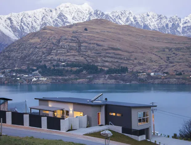 A contemporary single-storey house on a sloping lakeside site, with dark cladding, flat rooflines, large windows, and a sheltered outdoor terrace. The home overlooks a calm lake and a dramatic snow-capped mountain range, with a sparsely vegetated hillside and scattered settlement across the water.