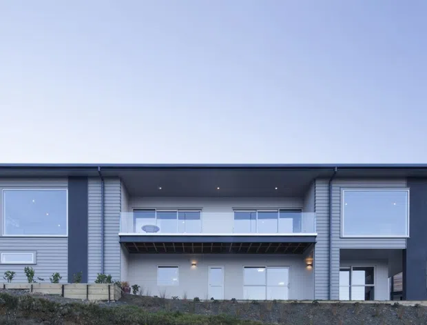 Front exterior view of a contemporary two-storey residence with a broad low-pitched roof, symmetrical grey horizontal cladding, dark structural framing, large square windows, and a recessed central balcony above a sheltered entry and glazed lower level. The house sits on a sloped site with wooded terrain in the background.