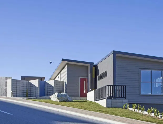 Street-level exterior view of a contemporary single-storey residential house on a sloping site. The home has grey horizontal weatherboard cladding, a low-pitched roof, large windows, a red front door, and a small raised entry porch with black metal balustrading. Concrete block boundary walls and a paved roadside verge are visible, with rugged mountain terrain in the distance.