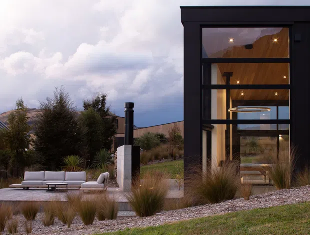 A contemporary dark-clad pavilion or extension viewed from a landscaped outdoor area. Floor-to-ceiling glazed openings reveal a warm timber-lined interior with recessed spotlights and a circular pendant or ceiling feature. In front is a paved terrace with a light outdoor sectional sofa, surrounded by ornamental grasses, shrubs, gravel, and low planting. A tall flue-like element and neighbouring low forms are visible in the background beneath a cloudy sky.