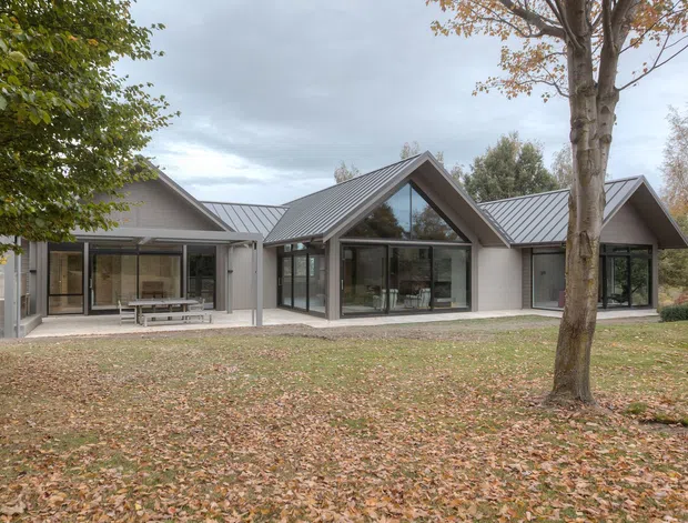 A low, modern residential building set in a landscaped garden, composed of several connected gabled volumes. The exterior features light rendered walls, standing-seam metal roofs, extensive dark-framed glazing, and a prominent glazed triangular gable. Mature deciduous trees and autumn leaves surround the house.