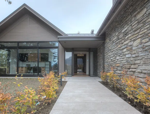 Exterior view of a contemporary single-storey residence with a steep gabled roof, extensive dark-framed glazing, and a prominent rough-stacked natural stone wall. A concrete pathway leads through landscaped planting to the recessed entry, with glimpses of the open-plan interior visible through the windows.