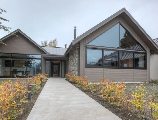 Single-storey contemporary gabled house viewed from the landscaped front approach. The exterior combines grey horizontal cladding, dark stone masonry, extensive triangular and rectangular glazing, and a standing-seam metal roof. A concrete path leads through low ornamental planting to the recessed entry, with interior spaces visible through the glazing.