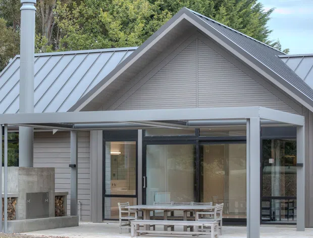 Exterior view of a contemporary gabled home with grey weatherboard cladding, standing-seam metal roofing, large glazed openings, and a covered outdoor entertaining area with a built-in fireplace and picnic-style dining table. Mature trees surround the house.
