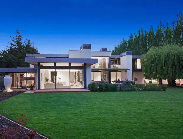 A large contemporary multi-level residence photographed at dusk from the landscaped front lawn. The house features crisp white and grey cubic forms, broad flat roof planes, extensive floor-to-ceiling glazing, projecting cantilevered sections, and illuminated interiors. Mature trees, manicured lawn, planting beds, and a curved paved garden path frame the property.