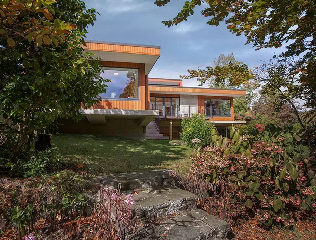 Exterior view of a modern, low-rise home set within a landscaped garden. The house features strong horizontal roof planes, extensive glazing, warm timber-toned cladding or trim, concrete elements, and a stepped stone garden path leading toward the entry.