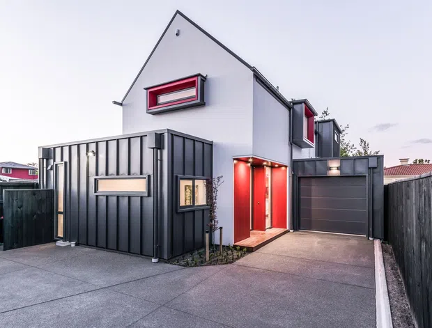 Contemporary two-storey residential house viewed from the driveway, featuring a steep gabled white-rendered upper volume, dark vertical metal-clad lower facade, red-framed windows and entry door, and an integrated dark garage. The paved driveway and timber boundary fencing frame the compact urban frontage.
