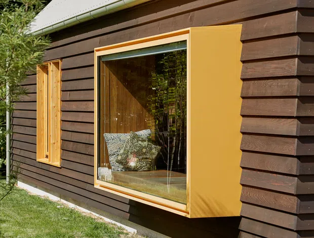 Close-up exterior view of a compact building clad in dark horizontal timber boards, with a projecting mustard-yellow window surround and timber-framed glazing. The window reveals a warm timber-lined interior with patterned seating, while surrounding greenery and lawn provide a natural setting. A light metal roof edge is visible above.