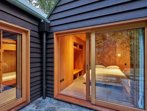 A close exterior view of a dark horizontal-clad building enclosing a small courtyard or paved outdoor area. Warm timber-framed glazing and sliding doors reveal a timber-lined bedroom with built-in shelving and a bed inside. The image highlights the contrast between dark exterior cladding, natural timber frames, and warm interior lighting.