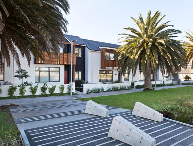Exterior view of a contemporary multi-unit residential building with white rendered walls, dark pitched metal roofs, extensive glazing, and warm timber balcony screens. The building fronts a landscaped communal lawn with mature palm trees, low planting, paved paths, and a modern slatted outdoor seating area in the foreground.