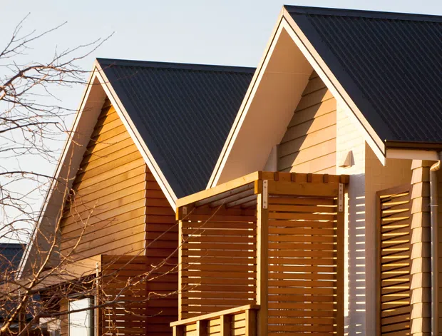 Exterior view of a contemporary timber-clad residential building with multiple steep front gables and dark corrugated metal roofing. Warm horizontal timber weatherboards and slatted timber privacy screens define the facade, with strong afternoon sunlight creating pronounced shadows.