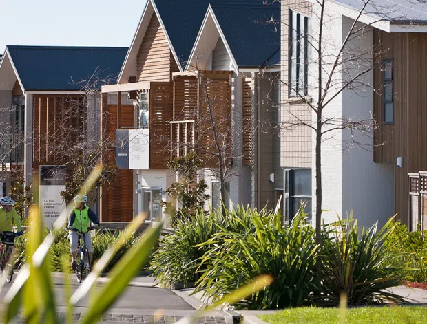 Exterior streetscape view of a contemporary multi-unit residential development, with repeated steep gabled roof forms, timber-clad facades, white rendered sections, balconies, and landscaped pedestrian paths. Cyclists are visible in the foreground, while ornamental grasses and shrubs soften the frontage.