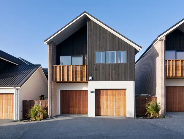 Exterior view of a row of compact multi-storey dwellings with steep gabled roofs, dark vertical timber-look cladding, pale rendered and brick walls, timber garage doors, and upper-level timber balconies. The homes face a paved shared driveway under clear daylight.