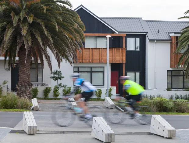 Street-level exterior view of a contemporary multi-unit residential building with dark vertical cladding, light rendered walls, pitched metal roofs, timber balcony screening, and large windows. Palm trees and landscaped planting frame the facade, while cyclists pass in the foreground.
