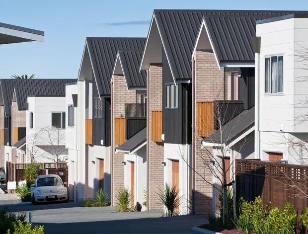 Exterior view of a contemporary multi-unit residential development on a sloping site. The row of narrow townhouses features repeated steep gabled dark metal roofs, pale brick and white rendered walls, dark vertical cladding, timber-toned balcony screens and fencing, and compact private driveways and planted front areas.