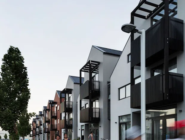 Street-level exterior view of a contemporary multi-storey residential development. A row of compact apartment or townhouse buildings lines a broad concrete footpath, with white and dark façades, black metal balconies, projecting pergola-like balcony frames, pitched metal roofs, and landscaped planting along the frontage. Pedestrians and parked cars are present in the streetscape, with a tree-lined urban or suburban setting.
