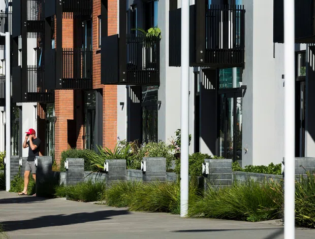Street-level view of a contemporary multi-storey residential development with repeated façades, projecting black metal balconies, vertical screening, exposed red-brick sections, pale rendered walls, and dark-framed glazing. Landscaped planter beds and a paved pedestrian forecourt line the building frontage.