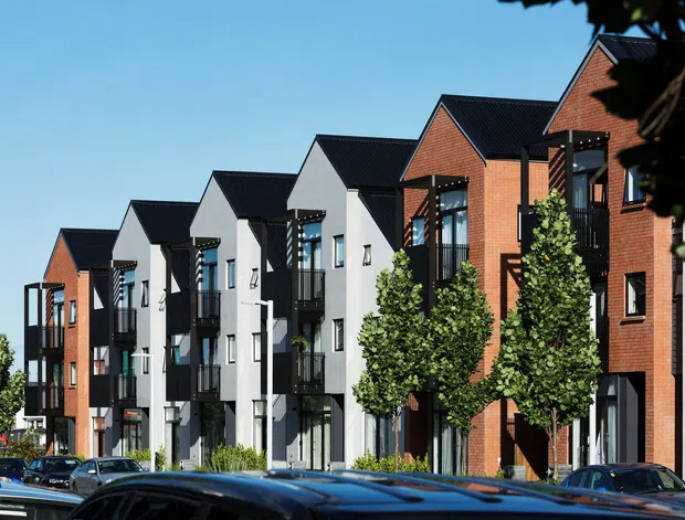 Exterior streetscape showing a row of contemporary multi-storey attached townhouses or apartment-like dwellings with repeated steep gabled roof forms, alternating red-brick and light-rendered facades, dark window frames, projecting balconies, and ground-level entrances. Mature street trees and parked cars occupy the foreground.