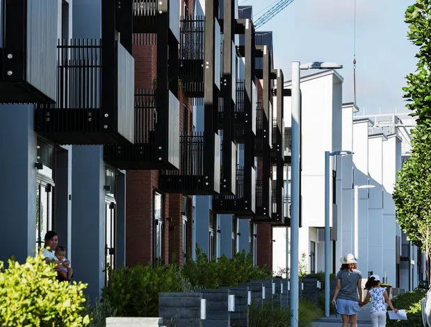 Street-level view of a contemporary multi-storey residential development with repeated projecting balconies, dark metal screens, exposed red brick sections, pale rendered walls, and landscaped pedestrian paths. The image looks along a suburban urban streetscape, with a construction crane visible in the background.