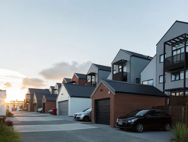 A row of contemporary multi-storey townhouses with attached garages lining a shared paved driveway. The homes combine dark vertical cladding, light rendered walls, brick garage-fronted volumes, pitched metal roofs, balconies, and pergola-like architectural screens. Several parked cars are visible in the driveway, with low planting at the foreground and warm low sunlight in the background.