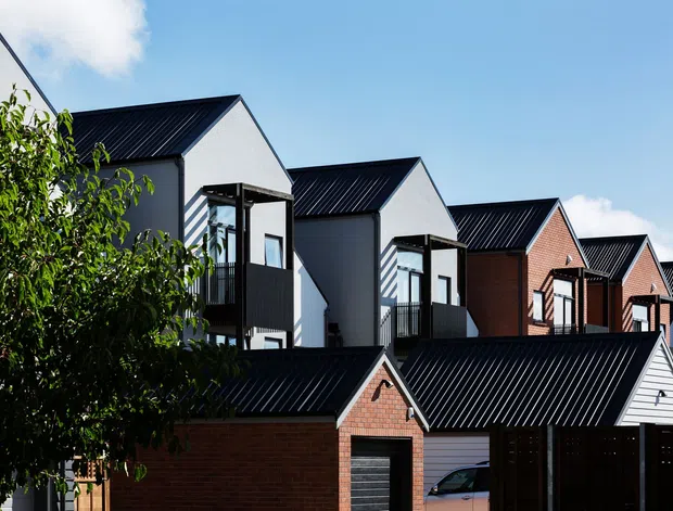 Exterior view of a row of contemporary attached homes with repeated steep gable roofs, dark metal roofing, brick and light-coloured cladding, and projecting upper-level balconies. Garages and privacy fencing are visible in the foreground, with mature planting partially screening the development.