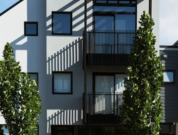 Front exterior view of a narrow, multi-storey contemporary townhouse with a steep gabled roof and white rendered facade. Black-framed windows, a dark glazed ground-floor entry, and two projecting balconies with vertical black metal balustrades and pergola-like overhead screens define the facade. Tall clipped trees and hedging frame the landscaped frontage, while strong sunlight casts graphic linear shadows across the building.
