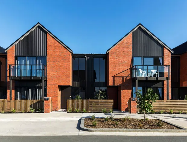 Street-facing view of a row of contemporary multi-storey attached residences. The homes feature steep gable forms, red brick walls, dark vertical metal cladding in the upper gable sections, black-framed glazing, balconies, and small fenced front gardens with timber screening. The image is captured in bright daylight against a clear blue sky.