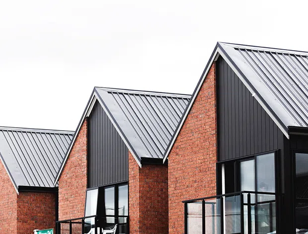 Exterior view of a row of attached or closely repeating modern residential units with steep pitched gable roofs, dark standing-seam metal roofing and cladding, red brick walls, large black-framed windows, and upper-level balconies. The repeated gable composition gives the building a townhouse-like form.
