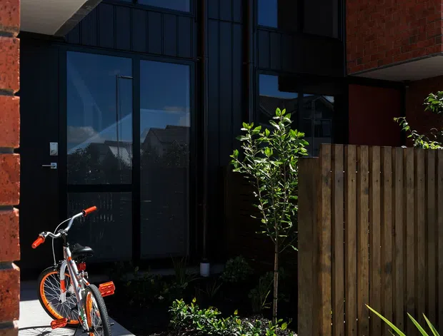 A narrow contemporary residential entrance courtyard framed by red brick walls and tall black-framed glazing. A black entry door opens onto a pale concrete path, with a timber privacy fence, landscaped garden beds, and a bicycle in the foreground. The facade features strong vertical proportions, reflective floor-to-ceiling windows, and dark metal framing.