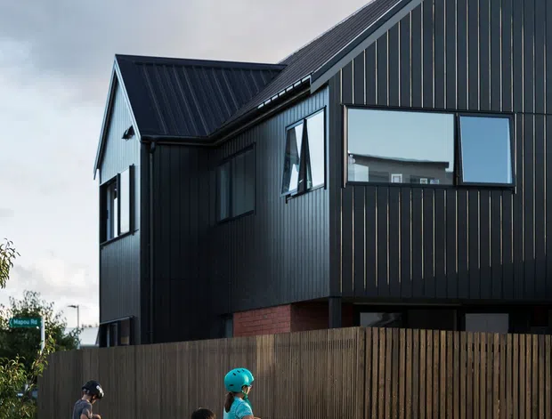 Exterior street-level view of a contemporary multi-storey house with steep gabled roofs, dark vertical cladding, large rectangular windows, and a timber paling fence. A small section of exposed red brick is visible at the lower facade. The home sits along a wet concrete footpath and driveway in a suburban setting.